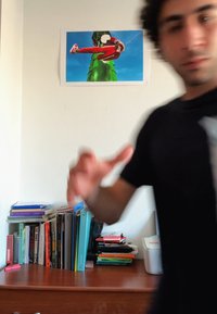Az íróasztalon különböző színű könyvek találhatók. A falon egy zöld szoborról készült poszter látható, amelyen egy piros ruhájú ember áll kék háttér előtt.