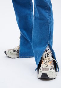 Jeans de mezclilla azul acampanados con aberturas laterales, combinados con zapatillas deportivas en crema y gris con paneles de malla y suelas negras.