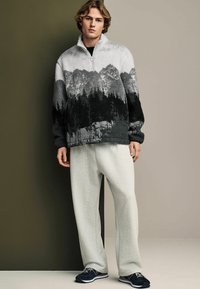 Polarowy pulower w kolorze szarym z czarnym wzorem leśnym, w połączeniu z jasnoszarymi spodniami dresowymi i granatowymi trampkami. Zamek na szyi, luźny krój.