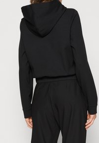 Persona vistiendo una sudadera negra con capucha y mangas largas, y pantalones negros a juego, vista desde atrás con las manos relajadas a los lados.