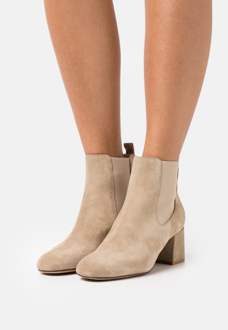 Bottes beige en daim à talons blocs portées sur jambes nues, présentées sur un fond clair uni.