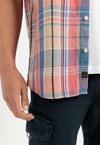 Korte mouwen geruit shirt in roze, blauw en beige met een afgeronde zoom en knoopdetails. Gecombineerd met een donkere broek met een klepzak.