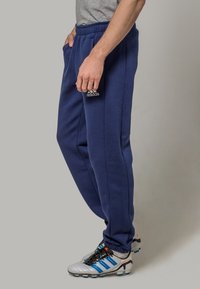 Mies, который одет в темно-синие спортивные штаны Adidas и белые футбольные бутсы Adidas, стоит на фоне без узоров.