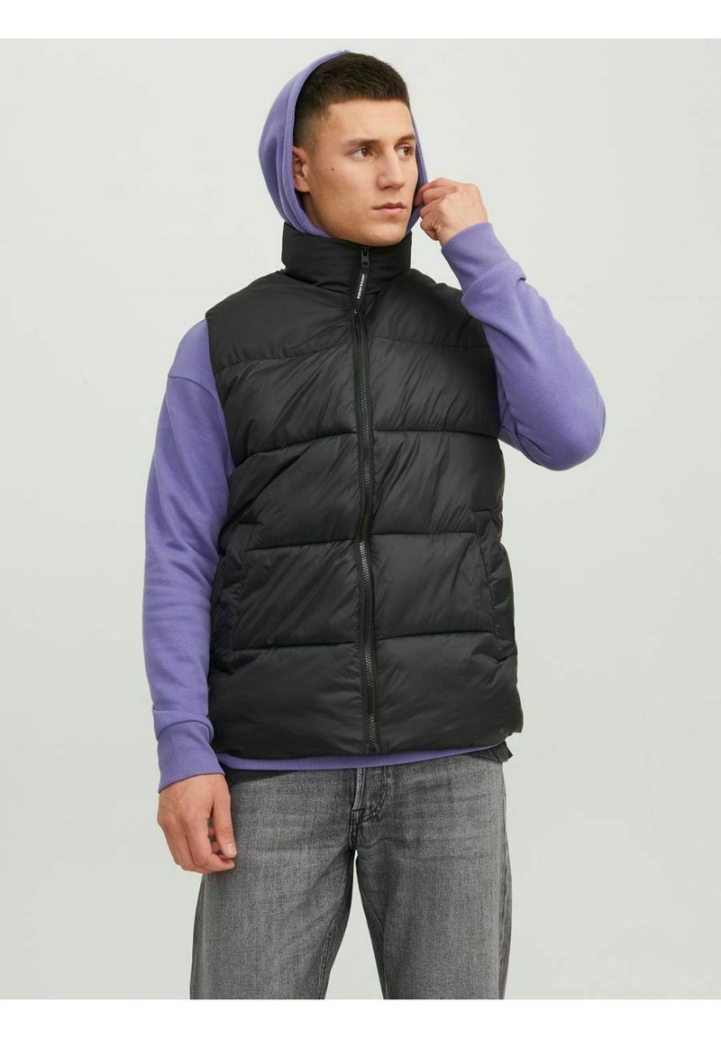Black puffer vest with a high collar, zip closure, and quilted design, worn over a purple long-sleeve shirt. Gray jeans complete the outfit.