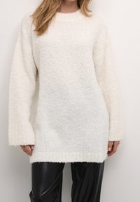 Pull en maille blanche avec un matériau texturé et doux. Il présente un col rond et des manches longues et larges. Associé à un pantalon noir brillant.