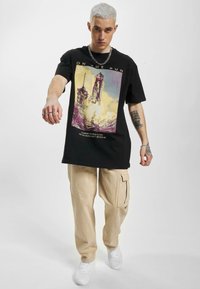 Sort grafisk t-shirt med et design af en raketopsendelse, kombineret med rummelige beige cargo-bukser og hvide sneakers. Intrikate tatoveringsdetaljer synlige.