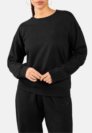 Frau trägt schwarzen Reebok-Sweatshirt und passende schwarze Hose, Hände vor dem Körper verschränkt, einfarbiger heller Hintergrund.