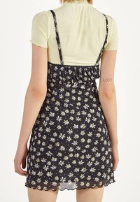 Svart klänning med blommönster av tusensköna, spagettiaxlar, volangliv och scalloped nederkant. Bärs över en ljusgul åtsittande topp.