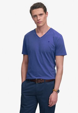 Hombre que lleva una camiseta azul de cuello en V, pantalones azul marino, cinturón marrón y reloj de pulsera color beige, de pie con una mano en el bolsillo sobre fondo blanco.