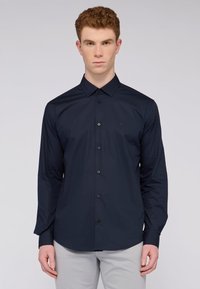 Camicia button-up blu navy con colletto a punta, maniche lunghe e bottoni neri. Presenta un piccolo logo sul lato sinistro del petto.