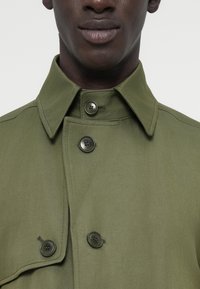 Nahaufnahme einer Person, die einen olivgrünen Mantel mit weitem Kragen und vier sichtbaren großen Knöpfen trägt, wobei ein Teil des Gesichts und des Halses zu sehen ist.