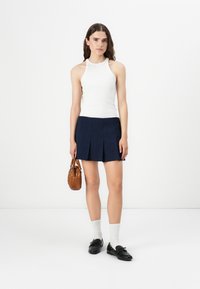 Weißes geripptes Tanktop, kombiniert mit einem marineblauen, plissierten Minirock, schwarzen Loafers und einer kleinen braunen Handtasche. Einfaches und klares Design.