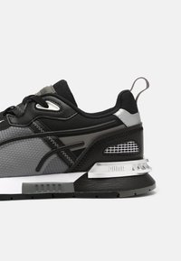 Baskets Puma noires et grises avec semelle rembourrée, dessus en mesh, languette au talon et surpiqûres décoratives sur fond blanc.