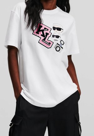 Osoba oblečená v bílém tričku s růžovými nápisy "Karl Lagerfeld", karikaturami obličejů se slunečními brýlemi a grafikou fotoaparátu, doplněná černými kalhotami.