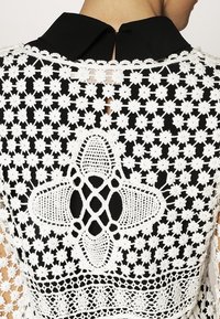 Spetsblus med svart krage, vita blommönster och en intrikat blommönster på svart bakgrund. Utsnitt på ryggen.