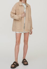 Veste en denim beige avec grandes poches à l'avant, fermetures à boutons et coupe ample, associée à un débardeur à motifs et un short beige. Sandales marron à enfiler.