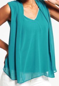 Haut sans manches teal en tissu léger et transparent, avec un design en v et un superposition ouverte dévalant vers le bas. Accents blancs visibles.