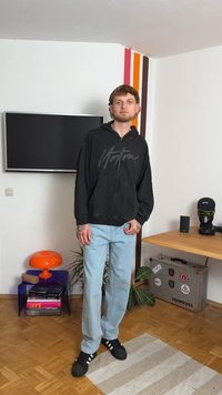 Crna zip-up hoodie s logotipom, svijetloplave traperice i crne tenisice s bijelim prugama. Unutarnje okruženje s namještajem i dekorom.