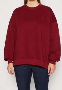 Sweatshirt oversize bordeaux avec un col rond, des poignets et un ourlet côtelés, arborant un logo brodé au niveau de la poitrine. Tissu en mélange de coton doux.