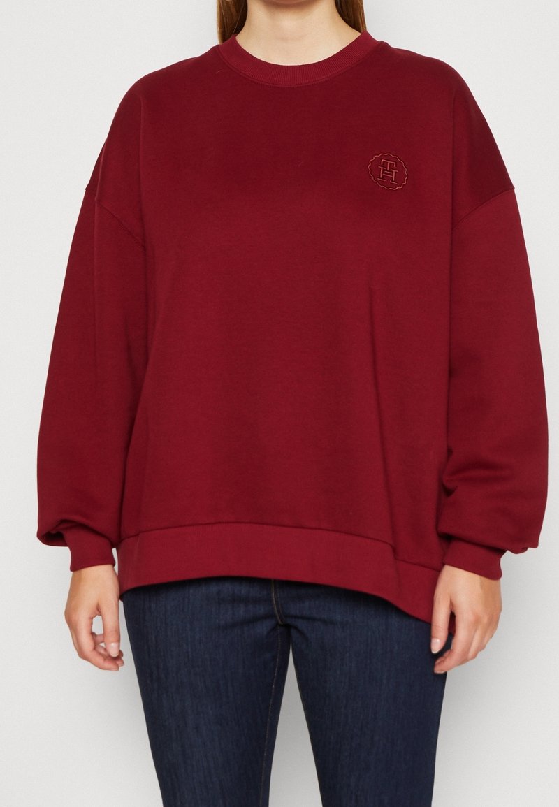 Sweatshirt oversize bordeaux avec un col rond, des poignets et un ourlet côtelés, arborant un logo brodé au niveau de la poitrine. Tissu en mélange de coton doux.
