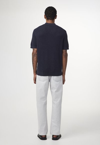 Tummansininen lyhythihainen polo-paita, jossa on teksturoitu neulottu muotoilu, yhdistettynä vaaleanharmaisiin suoriin farkkuihin ja mustiin litteäpohjaisiin kengiin.