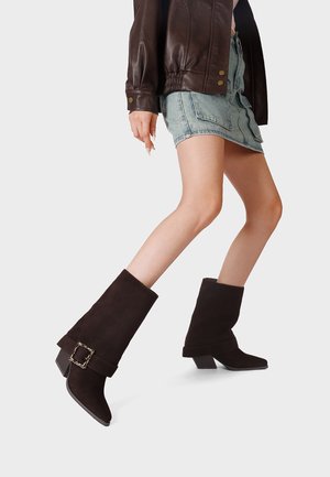 Persona che cammina indossando stivali in camoscio marrone scuro a metà polpaccio con dettaglio fibbia, minigonna di jeans e giacca di pelle marrone su sfondo chiaro.
