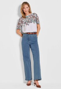 Weißes Oberteil mit buntem Paisley-Stickerei, kombiniert mit hellblauen weiten Jeans und einem braunen Gürtel. Braune Absatzschuhe runden den Look ab.