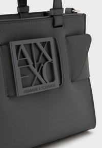 Melna ādas rokassoma ar kvadrāta Armani Exchange logo plāksni un izšūtām siksnas detaļām uz baltā fona.