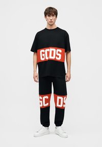 Ung mand iført sort oversized T-shirt og bukser med brede, røde, horisontale striber og hvide "GCDS"-logoer, kombineret med hvide sneakers.