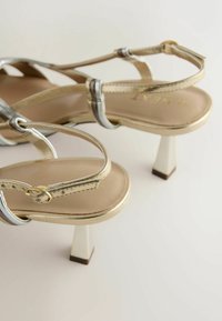 Sandales à talons dorées à brides avec talons carrés blocs et brides à boucle autour de la cheville, posées sur une surface claire.