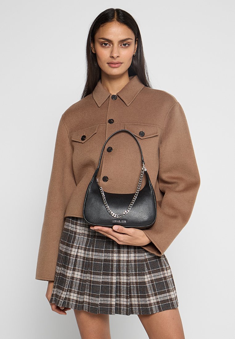 Borsa a tracolla in pelle nera con catena, forma curva e texture a granuli. Il modello indossa una giacca marrone e una gonna scozzese, mentre tiene la borsa.