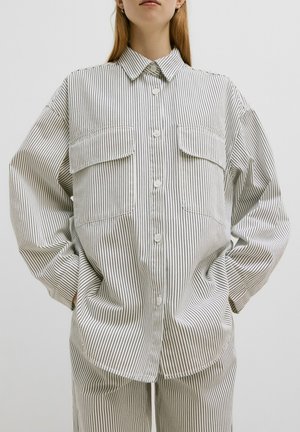 Chemise à rayures blanche et noire en coton, dotée de deux poches avant et d'une coupe décontractée avec des épaules tombantes.