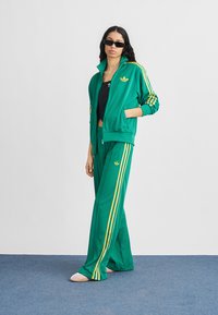 Γυναίκα με πράσινη φόρμα Adidas με κίτρινες ρίγες, μαύρο τοπ, γυαλιά ηλίου, στέκεται με το ένα χέρι στην τσέπη σε μπλε πάτωμα.