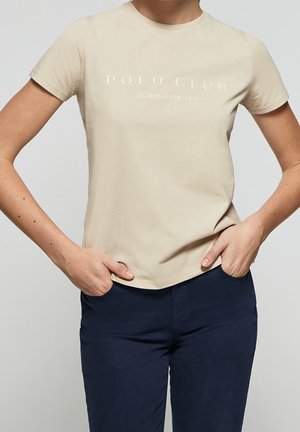 Beige lyhythihainen T-paita, jossa teksti "Polo Club Established 2012", pukeutuneena henkilö, jolla kädet taskuissa, yhdistetty tummansinisiin housuihin.