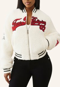 Biała, miękka kurtka bomber z czerwonym haftowanym logo i akcentami, czarne prążkowane mankiety i pas, zamek błyskawiczny z przodu, boczne kieszenie.