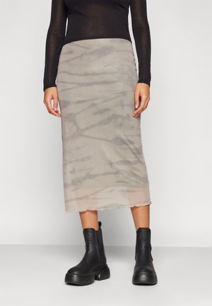 Personne portant une jupe midi beige transparente, un haut à manches longues noir et des bottines noires épaisses, debout devant un fond uni.