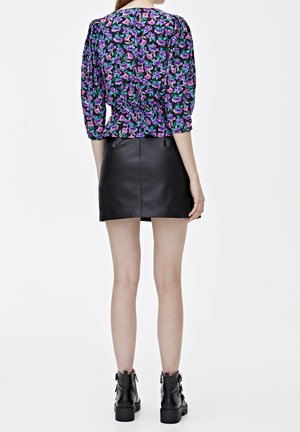 Femme portant un chemisier violet à fleurs avec des manches trois-quarts, une mini-jupe en cuir noir et des bottines noires lacées, debout de dos.