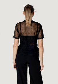 Blusa negra transparente con mangas cortas, con un patrón sutil. La vista trasera revela transparencia y un corte relajado. Combinada con pantalones negros.