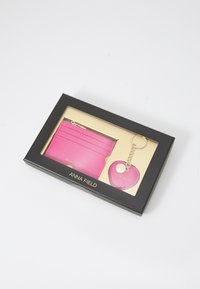 Pink wallet and heart-shaped keychain set in a black box with gold accents. Wallet has card slots and smooth texture. Keychain features a logo.