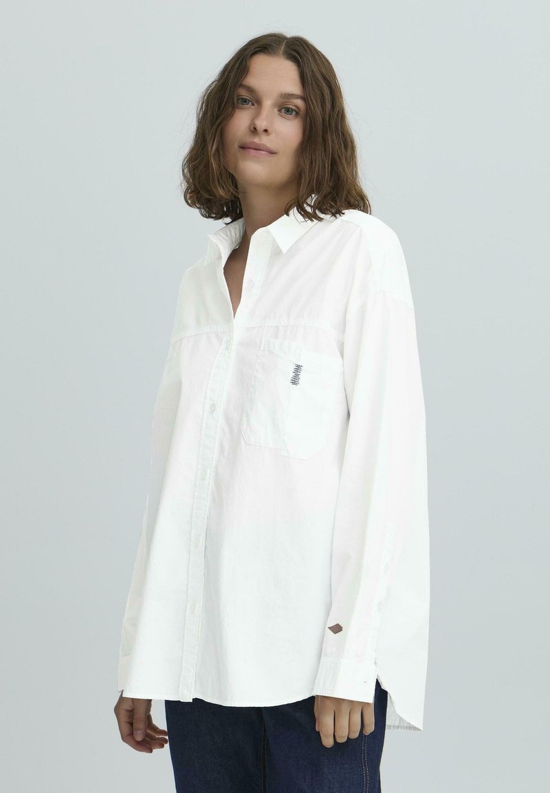 Balta medvilninė ilgomis rankovėmis palaidinė su mygtukų apykakle, krūtinės kišenių ir subtiliomis siuvinėjimo detalėmis. Vilkima su tamsiomis džinsinėmis kelnėmis.