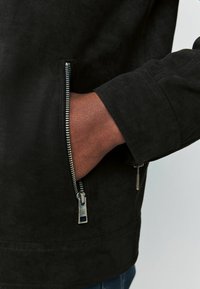 Sort ruskindjakke med lynlåsdetaile ved lommen. Stoffet har en glat tekstur, og designet har en tætsiddende silhuet.