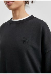 Felpa nera con scollo a girocollo, polsini a coste e un discreto logo ricamato sul petto. Tessuto morbido e texturizzato.