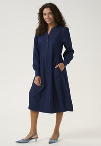 Marinblå denimklänning med långa ärmar, knapplist, sidofickor och utsvängd fåll. Matchad med ljusblå slingbacks.