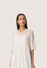 White, textured dress featuring a V-neckline, short puff sleeves, and gathered bodice. The fabric has a subtle dotted pattern throughout.