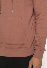 Felpa marrone chiaro con tasca a marsupio, cappuccio con cordino, polsini e orlo a coste. Tessuto morbido e testurizzato con cuciture visibili.