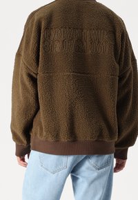 Brązowy polarowy sweter z teksturowaną powierzchnią, z wytłoczonym logo z tyłu i ściągaczami przy rękawach. Połączony z jasnoniebieskimi dżinsami.