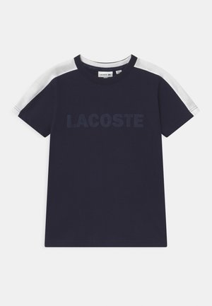Granatowy t-shirt z krótkim rękawem, z białymi paskami na ramionach i napisem "LACOSTE" na piersi w lekko ciemniejszym odcieniu.