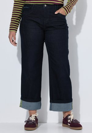 Person iført mørke vide jeans med opbukket kant nederst, en langærmet stribet skjorte og bordeaux sneakers med hvide snørebånd.