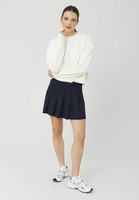 Jeune femme aux cheveux foncés portant un pull blanc, une jupe plissée marine et des baskets blanches, debout avec la main près du menton sur un fond uni.