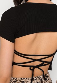 Černý žebrovaný crop top s vykrojeným zadním dílem, s překříženými ramínky uvázanými v pase. Krátké rukávy a hladký povrch.